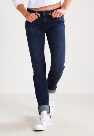 Vaqueros azul oscuro con un corte recto, confeccionados en denim. Pliegues en los puños, combinados con zapatillas blancas para un look casual.