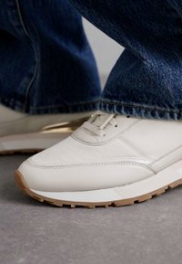 Sneaker bianca in pelle con punta arrotondata, suola in gomma marrone chiaro e dettagli di cucitura sottili. Abbinata a jeans in denim blu.