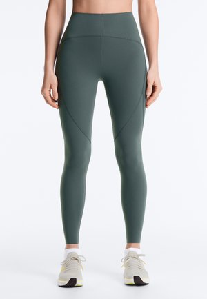 Predný pohľad na osobu nosiacu tmavozelené legíny s vysokým pásom a zakrivenými švami, obutú v bielych teniskách, na jednoduchom pozadí.