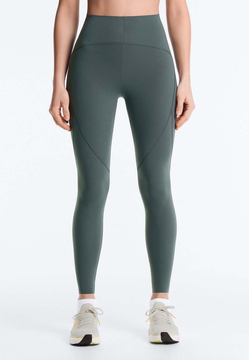 Vue de face d'une personne portant des leggings taille haute vert foncé avec des détails de couture courbés et des baskets blanches sur un fond uni.