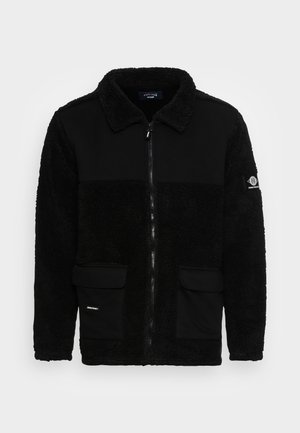 Sort jakke med fleece-materiale, lynlås foran, krave, patchlommer og en struktureret krop med glatte paneler.