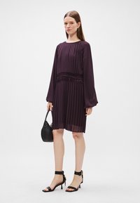 Fialové plisované šaty s dlhými rukávmi a smockovaným pásom, doplnené o čierne sandále na vysokom opätku a malú čiernu kabelku.