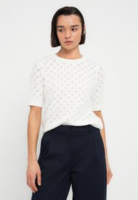 Λευκό πλεκτό πουλόβερ με κοντά μανίκια, που διαθέτει διαμαντένιο σχέδιο και στρογγυλή λαιμόκοψη, σε συνδυασμό με μπλε ναυτικό παντελόνι.