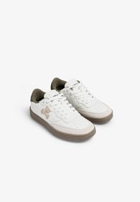 Zapatillas bajas blancas con acentos de gamuza beige, suelas marrón claro, logo de calavera y tibias en el costado, y forro interior verde.