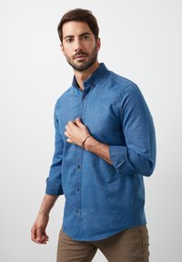 Camisa azul de botones hecha de tela texturizada, con cuello tipo camisa, mangas enrolladas y botones marrones, combinada con pantalones beiges.