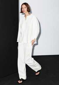 Weiße Leinenbluse mit lockerem Schnitt und fünf Knöpfen, kombiniert mit passenden Leinenhosen und schwarzen Sandalen. Glatte Textur, schlichtes Design.