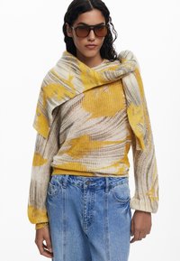 Gebreide trui met abstract patroon in geel en beige, rond de schouders gewikkeld, gecombineerd met high-waisted blauwe spijkerbroek.