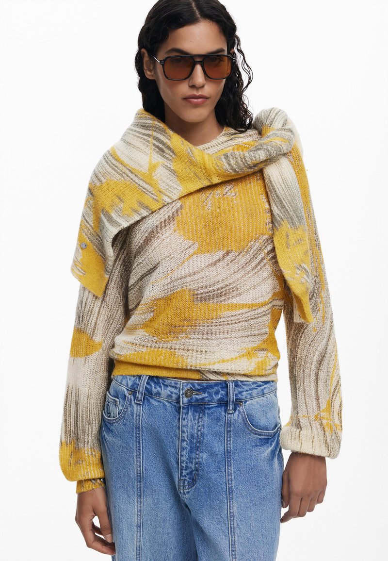 Gebreide trui met abstract patroon in geel en beige, rond de schouders gewikkeld, gecombineerd met high-waisted blauwe spijkerbroek.