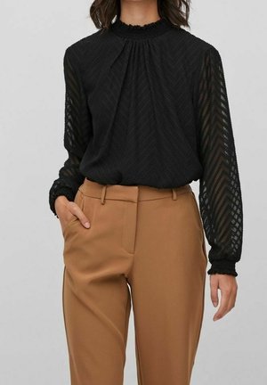Kobieta ubrana w czarną, teksturowaną bluzkę z długim rękawem z mankietami oraz beżowe, wysokowalne spodnie, z jedną ręką w kieszeni.