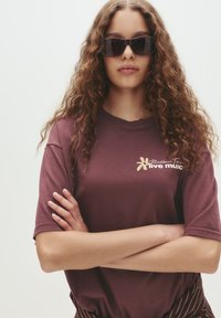 Ung person med langt krøllet hår iført mørke solbriller og en maroon "live music" t-shirt, med armene krydset mod en ensfarvet baggrund.