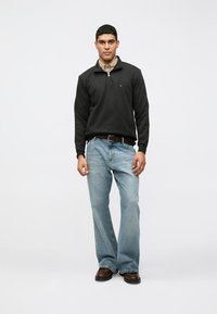 Felpa nera con zip, camicia colletto grigia chiara sotto, jeans dritti blu chiaro, scarpe marroni, in piedi contro uno sfondo neutro.