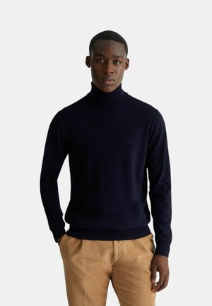 Maglione a collo alto blu navy realizzato in un tessuto liscio, con maniche lunghe e un design aderente. Abbinato a pantaloni in velluto a coste color cammello.