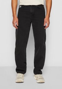 Black denim jeans with a straight cut, featuring front pockets and a branded patch on the waistband. Fabric has a slight texture.