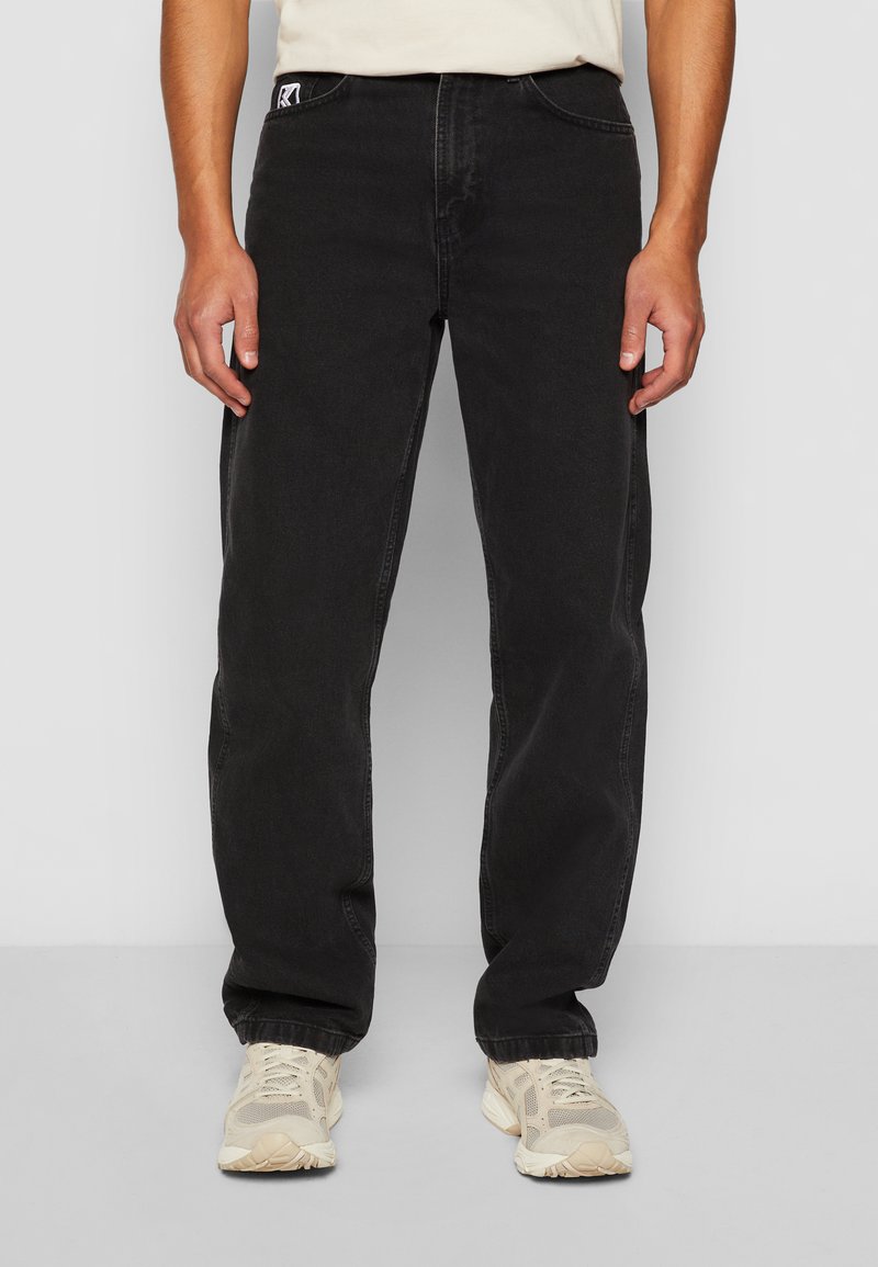 Black denim jeans with a straight cut, featuring front pockets and a branded patch on the waistband. Fabric has a slight texture.