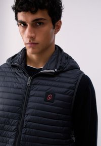 Jeune homme aux cheveux courts et bouclés portant une veste matelassée noire zippée par-dessus une chemise noire à manches longues, sur un fond uni.