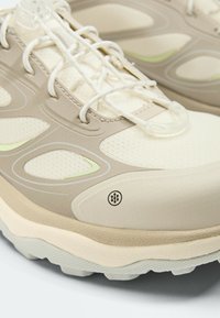 Gros plan d'une basket beige et crème avec tissu en mesh, surcouches en plastique, semelle épaisse et lacets blancs avec un verrouillage transparent.