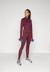 Burgundowy zestaw sportowy z długimi rękawami i obcisłymi legginsami, akcentowany różowymi szwami. Model trzyma niebieskie hantelki, nosząc szare trampki.