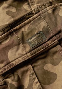 Camouflage-stof met een patchlabel met "Superdry" branding. Aardse tinten met zichtbare stiksels en een zachte textuur.