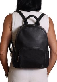 Persona che indossa un top bianco senza maniche e pantaloni, con uno zaino in pelle nera dotato di tasche con zip frontali e laterali.
