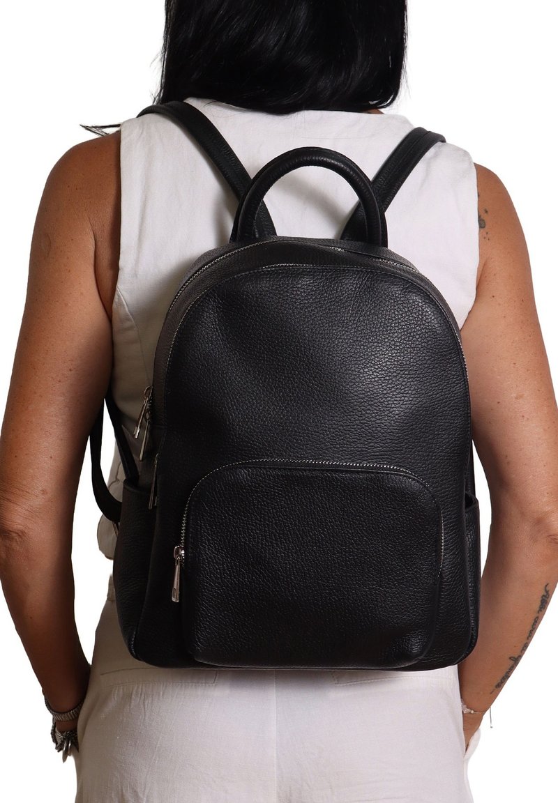 Persona che indossa un top bianco senza maniche e pantaloni, con uno zaino in pelle nera dotato di tasche con zip frontali e laterali.