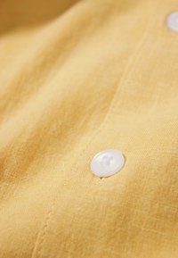 Primo piano di un bottone bianco cucito su un tessuto giallo testurizzato con cuciture visibili lungo la cucitura.