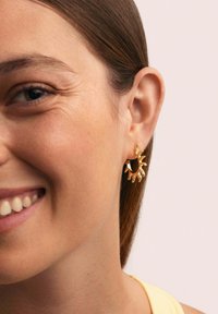 Orecchini a cerchio dorati con design a raggiera, caratterizzati da accenti testurizzati. L'orecchino pende da un piccolo chiodo d'oro sull'orecchio di un modello.