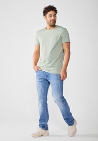 Camiseta de algodón verde claro con cuello redondo, acento de logo amarillo en el pecho, combinada con pantalones vaqueros de mezclilla azul y zapatillas beige.