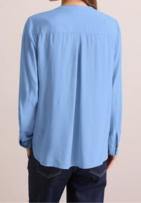Blouse bleu clair à manches longues avec un col rond, texture lisse et un pli central dans le dos, associée à un jean foncé.