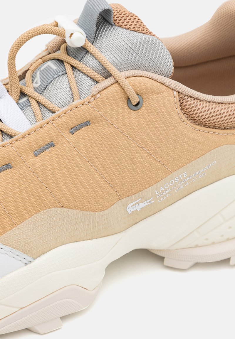 Baskets Lacoste avec une tige en tissu beige, des accents en maille, des détails gris et une semelle blanche texturée. Comprend un système de laçage léger.