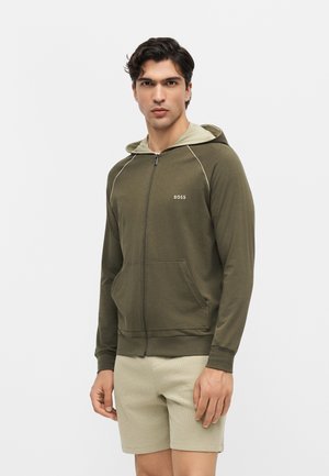 Mężczyzna ubrany w oliwkowo-zieloną bluzę z kapturem na suwak z logo „BOSS”, beżową podszewką kaptura i beżowe spodenki, stojący na jednolitym jasnym tle.