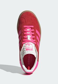 Czerwony zamszowy sneaker z różowymi akcentami, z trzema paskami, białym, wzorzystym językiem i beżową gumową podeszwą. Logo umieszczone na języku.