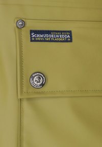 Zielono-oliwkowy wodoodporny materiał z przeszywaną klapą, z niebieską łatą z tkaniny i okrągłym czarnym guzikiem z białym tekstem.