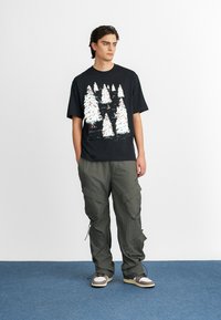 Must have mustaari t-särk koos valge männitüübi graafikaga, kombineeritud oliiviroheliste cargo-pükste ja beežide tossudega. Sile puuvillane tekstuur, mugav lõige.