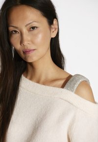 Off-the-shoulder crèmekleurige trui met een geribbelde halslijn en een metallic versierde band op één schouder. Zacht, gestructureerd materiaal.