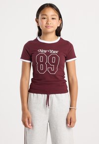 Ragazza con lunghi capelli scuri che indossa una maglietta maroon con la scritta New York 89 e pantaloni della tuta grigi chiari, in piedi davanti a uno sfondo bianco uniforme.