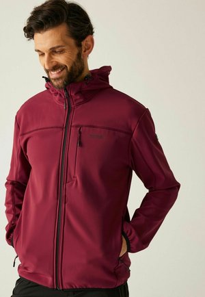 Mężczyzna w bordowej kurtce z kapturem, z suwakiem na piersi, ręce w bocznych kieszeniach, uśmiechający się i patrzący w dół na jednolitym tle.