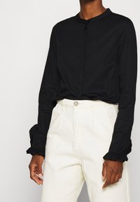 Camicetta nera a maniche lunghe con scollatura rotonda, dotata di chiusure a bottoni e polsini a volant, abbinata a pantaloni bianchi a vita alta.