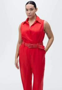 Combination rouge avec un col, taille ceinturée et jambes larges, fabriquée dans un tissu léger à la texture lisse.