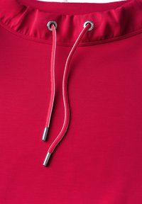 Roter Sweatshirt-Stoff mit gerafftem Halsausschnitt, ausgestattet mit silbernen Ösen und kontrastierenden roten Kordeln. Glatte Textur und einfaches Design.