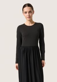 Black long-sleeve dress with a fitted top and pleated skirt. Smooth fabric at the top transitions to textured pleats at the bottom.