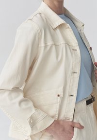 Krémová džínová bunda s knoflíkovým zapínáním, klasickým límcem, dvěma náprsními kapsami a kontrastním šitím. Nositá přes světle modré tričko.