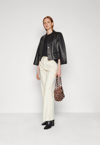 Schwarze Leder-Cropped-Jacke, enger schwarzer Oberteil, cremefarbene Weitbein-Hosen und schwarze Stiefeletten. Hält eine braune Kette-Taschen.
