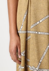 Beige textúrált ruha fekete és fehér márkás szalag részletekkel. Részletfotó egy kézről, amely a szövetet fogja, kiemelve a mintázatot.
