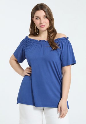 Frau trägt ein blaues, schulterfreies Kurzarm-Top und weiße Hose, posiert mit einer Hand an der Hüfte vor einem einfarbigen Hintergrund.