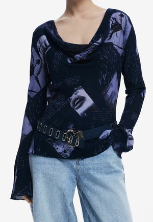 Femme portant un haut noir à manches longues avec des imprimés graphiques violets et un jean bleu clair, accessoirisée d'une ceinture noire avec œillets métalliques et boucle.