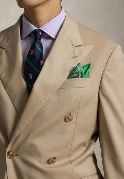 Giacca da abito beige doppiopetto con fazzoletto da taschino verde a fantasia, cravatta blu navy con righe verdi ed emblemi a scudo e camicia lilla chiaro.