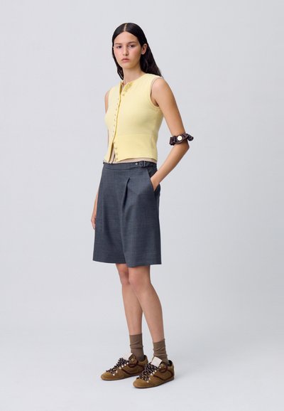 Top giallo lavorato a maglia senza maniche con dettaglio di bottoni, abbinato a pantaloni corti grigi a pieghe. Sneakers marroni e bianche con calzini marroni a motivi.