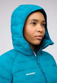 Teal puffjacka med huva, med en quiltad textur och framsidan dragkedja. Vit "MAMMUT"-logotyp synlig på vänster bröst.