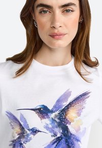 Maglietta bianca con una stampa ad acquerello di due colibrì nelle tonalità di blu e viola, arricchita da accenti luccicanti sulle ali.