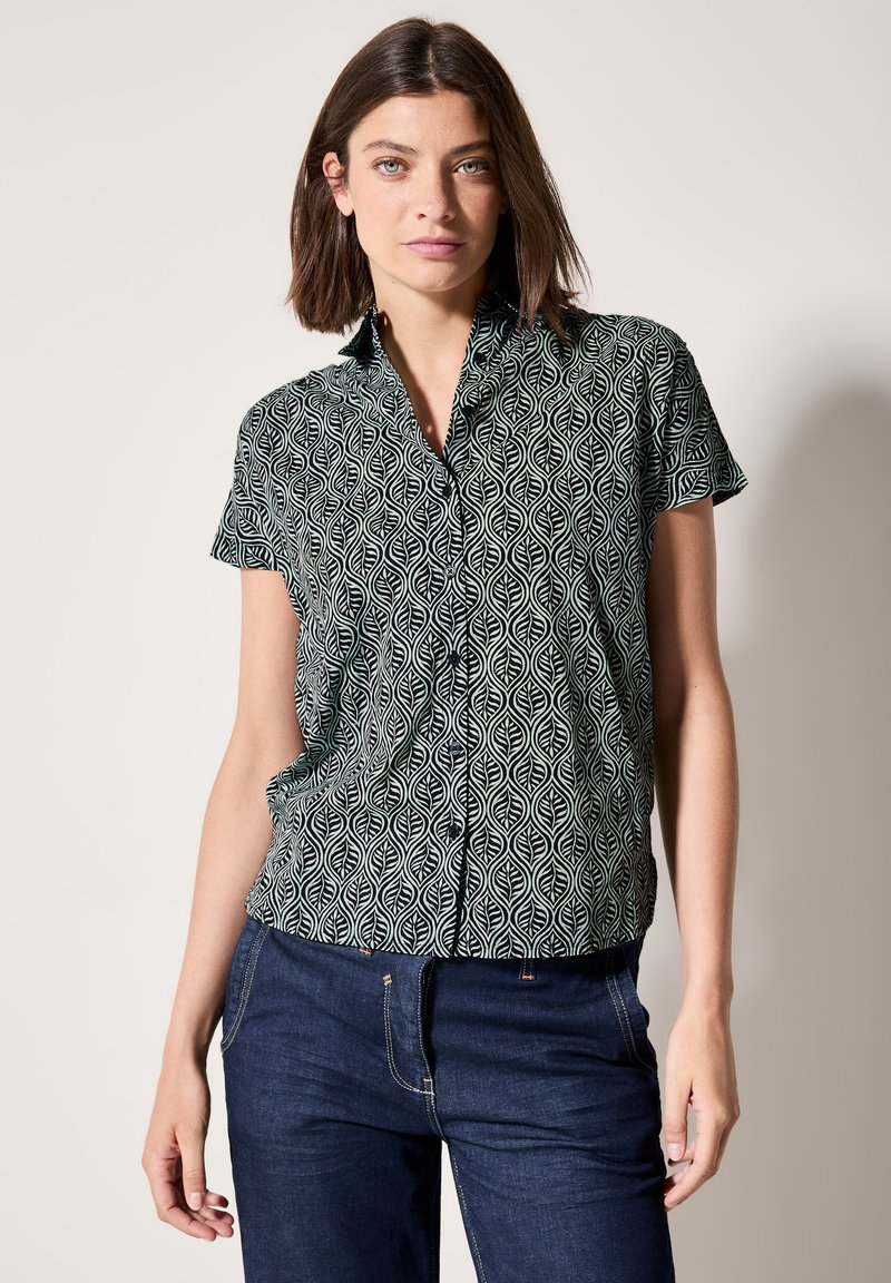 Camicia a maniche corte bianca e nera con motivo a foglie, colletto a punta e vestibilità rilassata. Abbinata a jeans scuri.